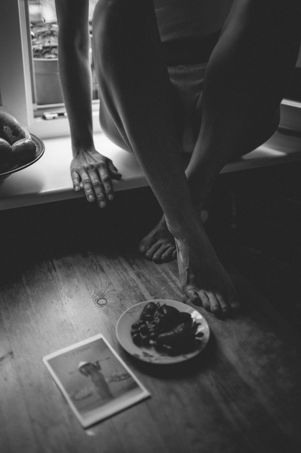 Moody black and white photograph showing bare feet, a plate of food, and a photograph on a wooden floor.