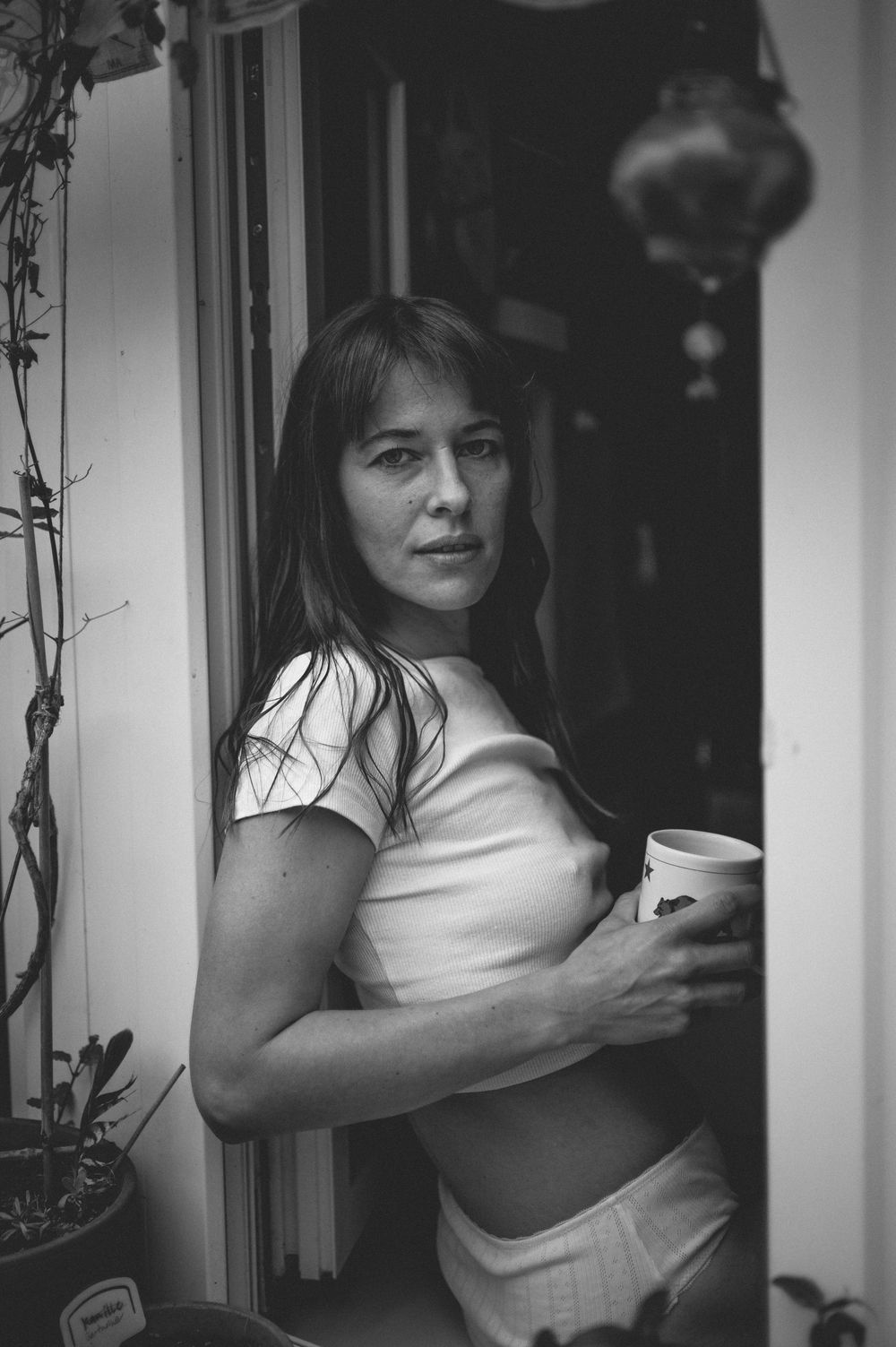 Black and white portrait shot of someone in a cropped top standing in a doorway holding a cup.