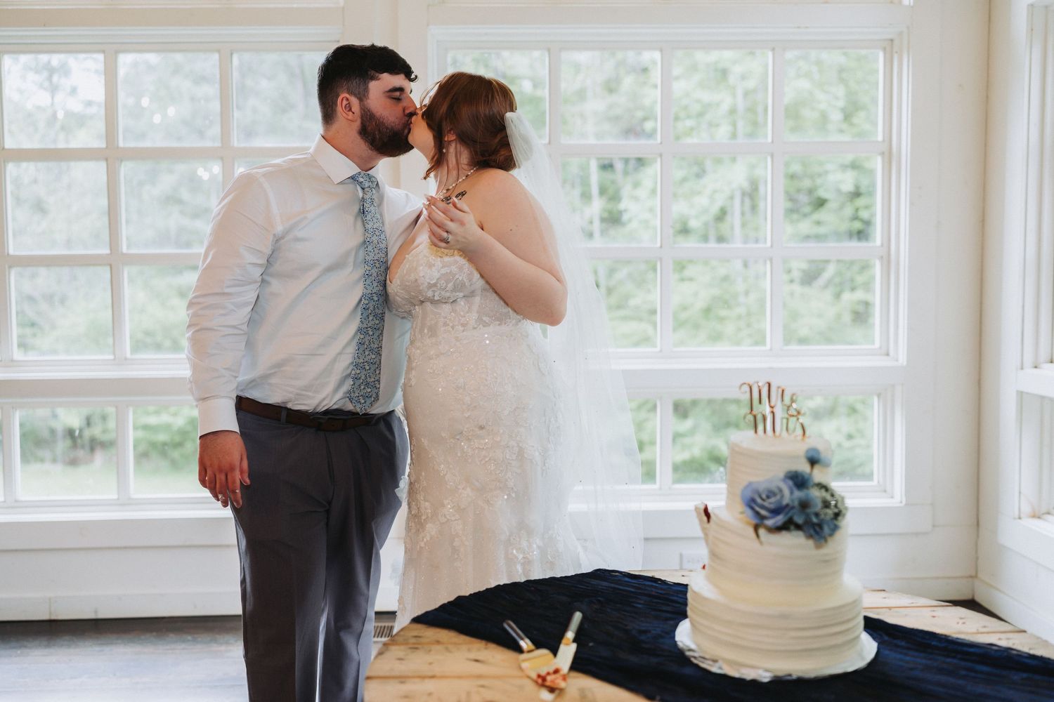 A bride and groom stand together in front of large windows with their wedding cake displayed on a table nearby.