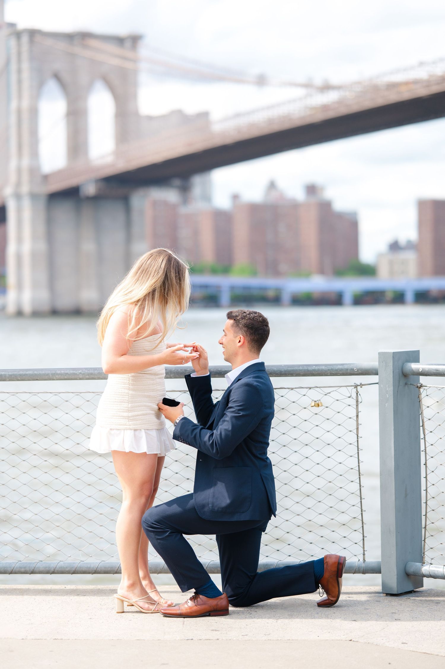 A romantic proposal moment near Brooklyn Bridge of a couple on a waterfront walkway during a sunny day captured by NYC proposal photographer