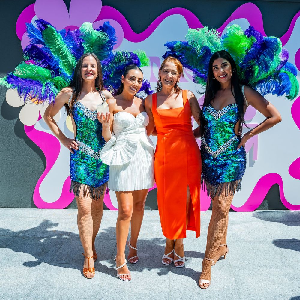 Four people in colorful cocktail dresses pose against a vibrant pink and white wall with turquoise feather decor.