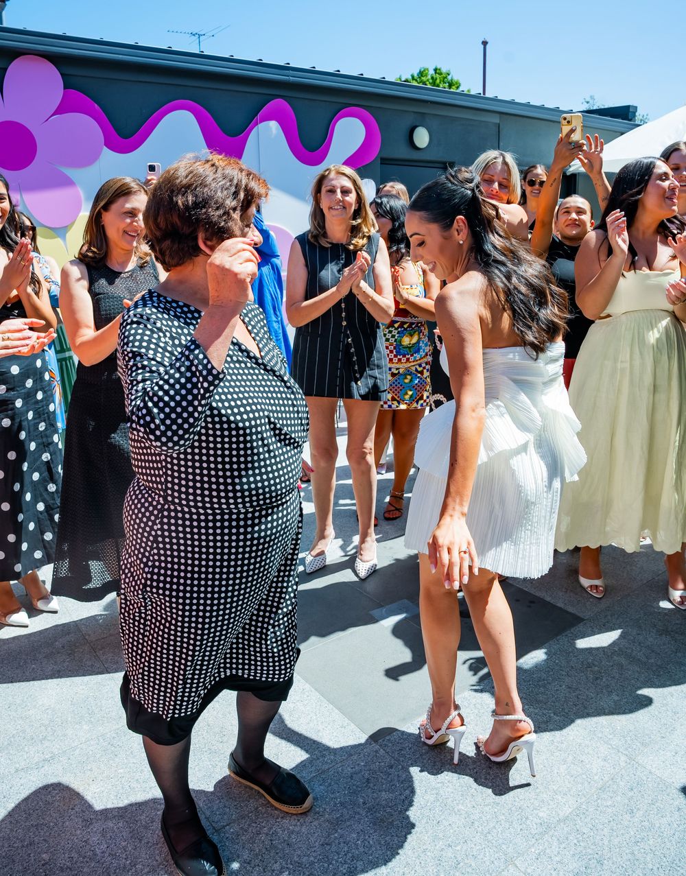 A lively outdoor celebration with a group of people dancing and socializing against a wall with colorful wavy patterns.
