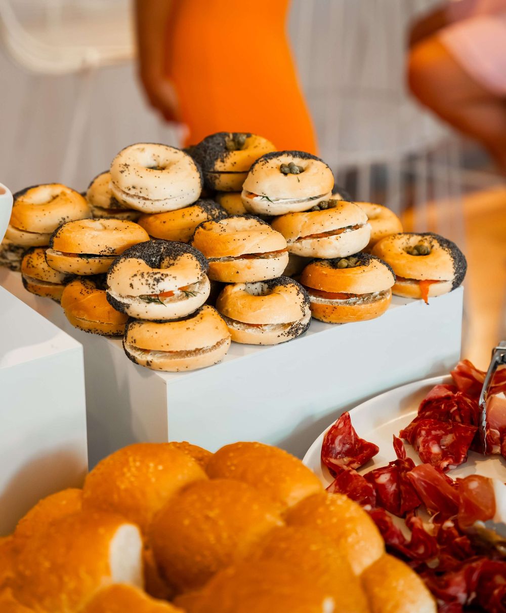 A platter of stacked black and white bagels with cream cheese filling displayed next to fresh oranges and pomegranate.