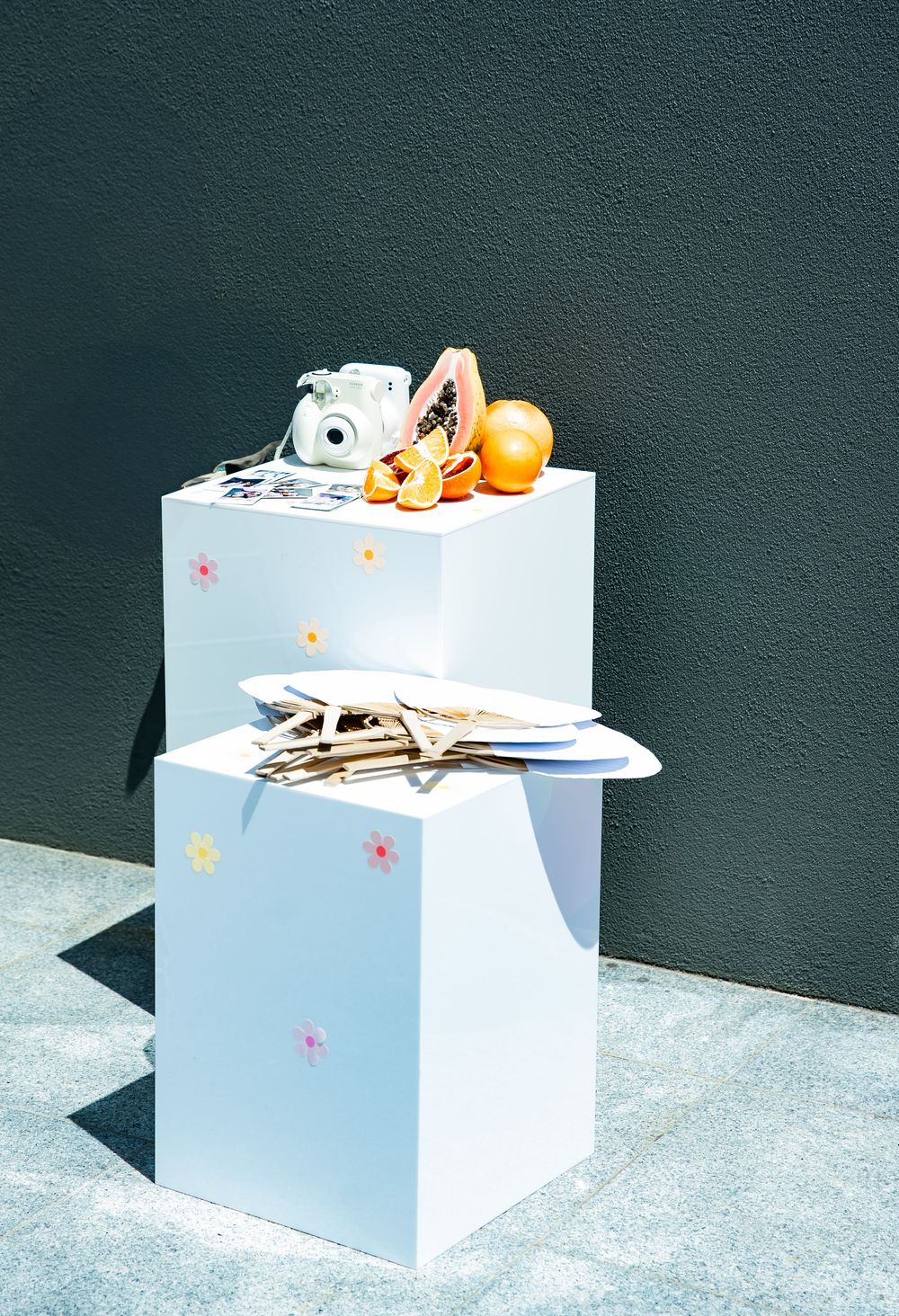 White vintage camera and oranges arranged on a light blue display pedestal with colorful star decorations against dark wall.