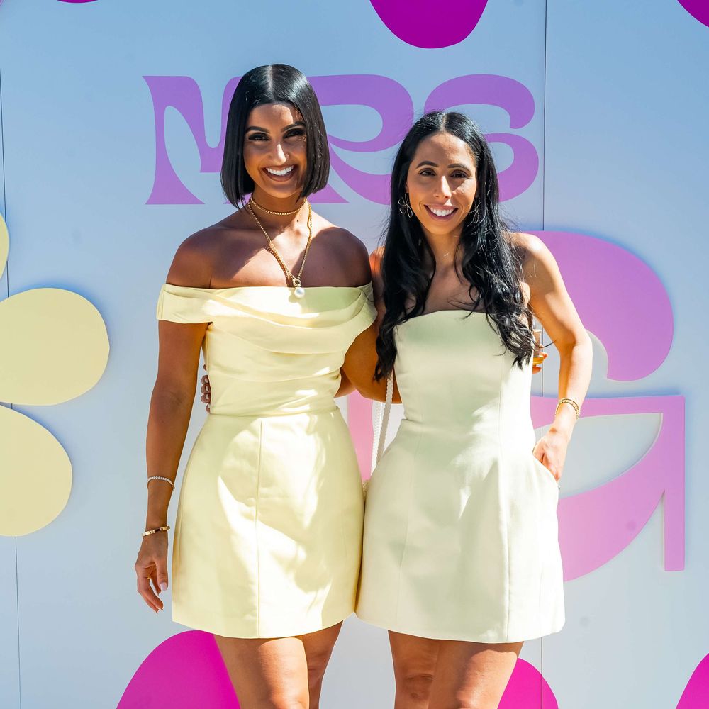 Two people stand together wearing yellow and light-colored summer dresses against a pastel backdrop with pink circles.