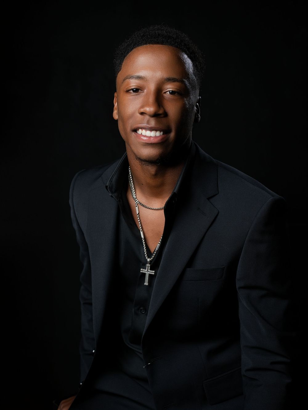 Young man in black suit and cross necklace smiling against dark background in professional portrait photo.