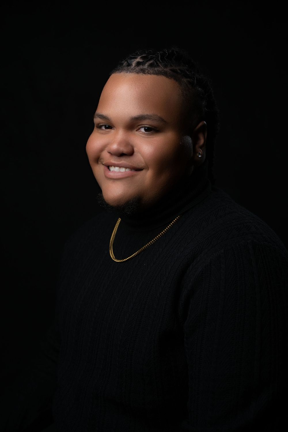 A portrait of a person wearing a black sweater and gold necklace against a dark background, with soft dramatic lighting.