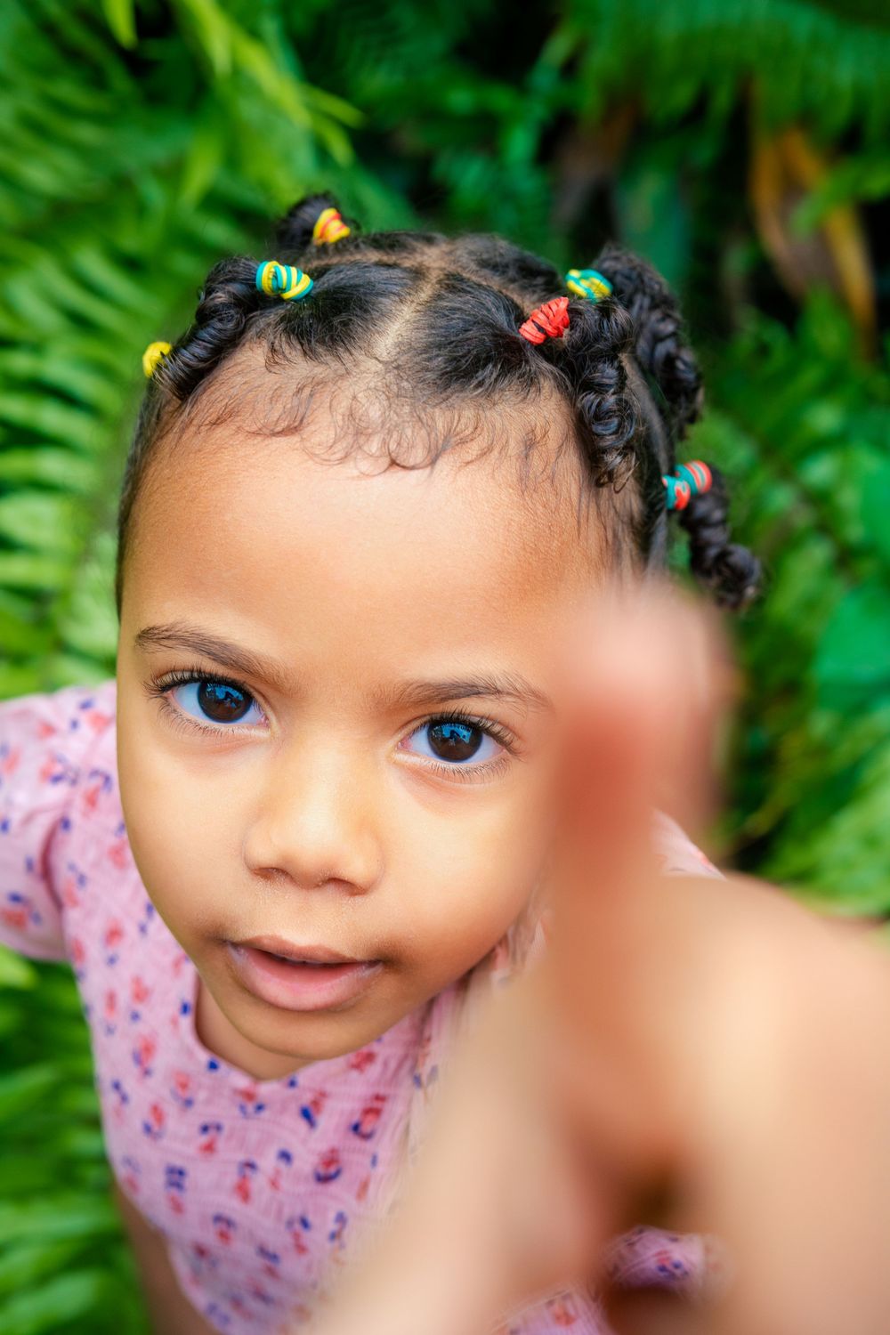 Portrait photo against green foliage background showing braided hair with colorful beads and pink floral pattern dress.