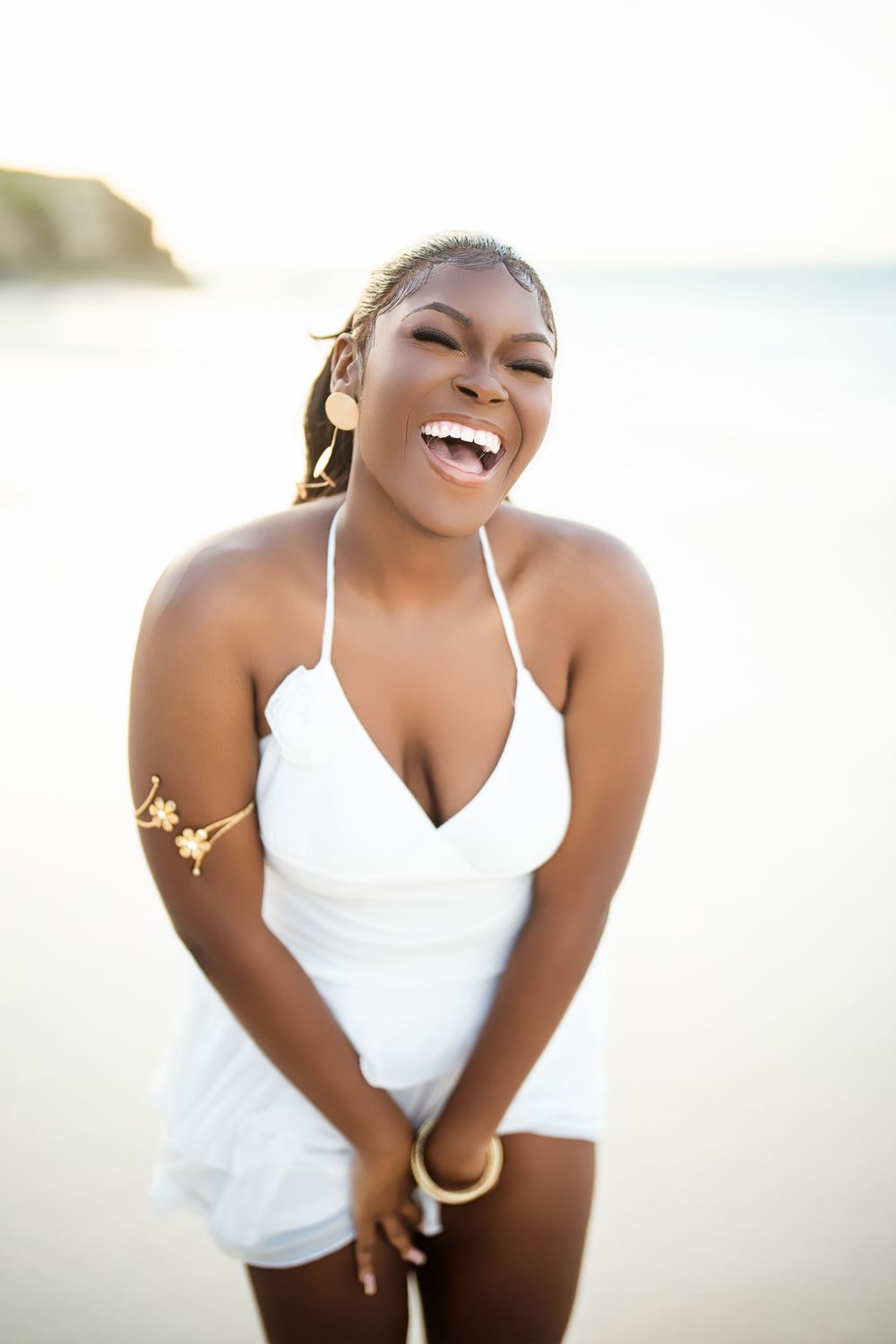 A joyful person in a white sundress laughs on a bright, sunny beach, wearing an arm cuff accessory.