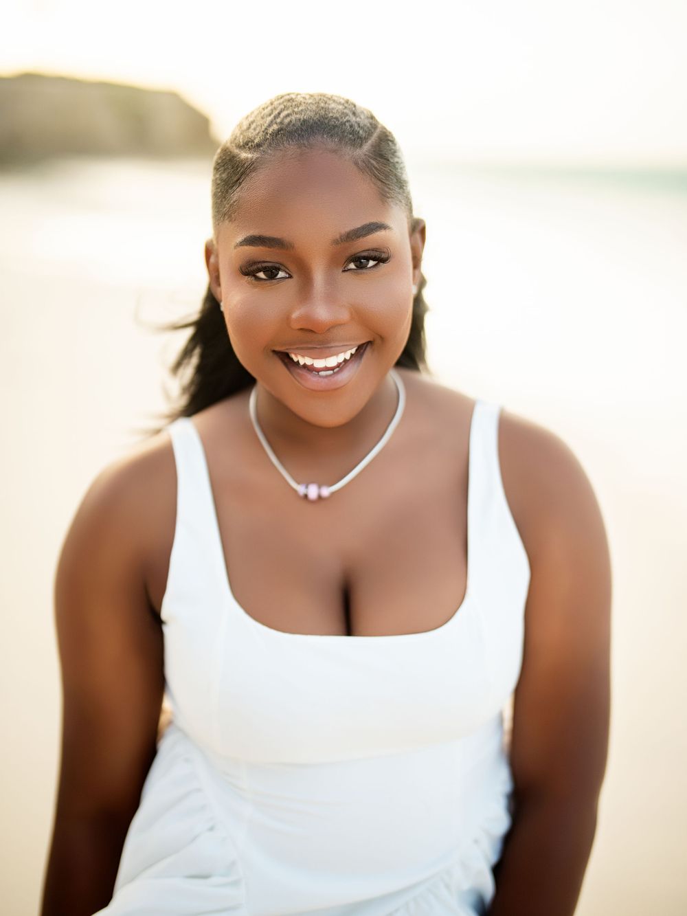 Portrait of someone wearing a white tank top and necklace smiling warmly at the beach during golden hour.