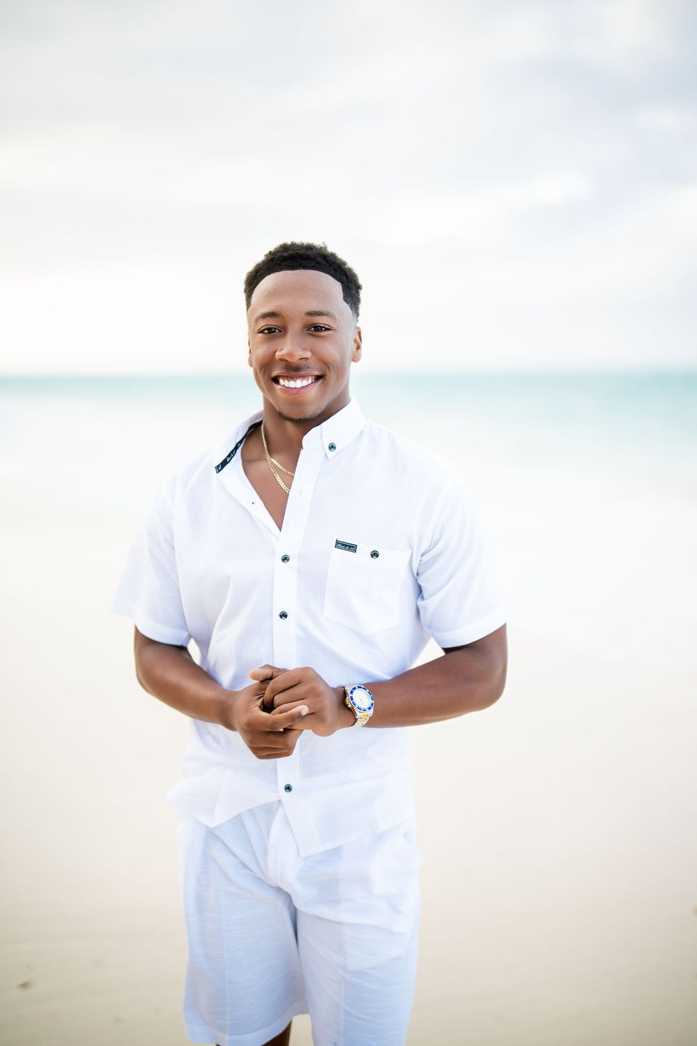 Smiling young Black man in white shirt and shorts standing on a tropical beach with turquoise water behind him.