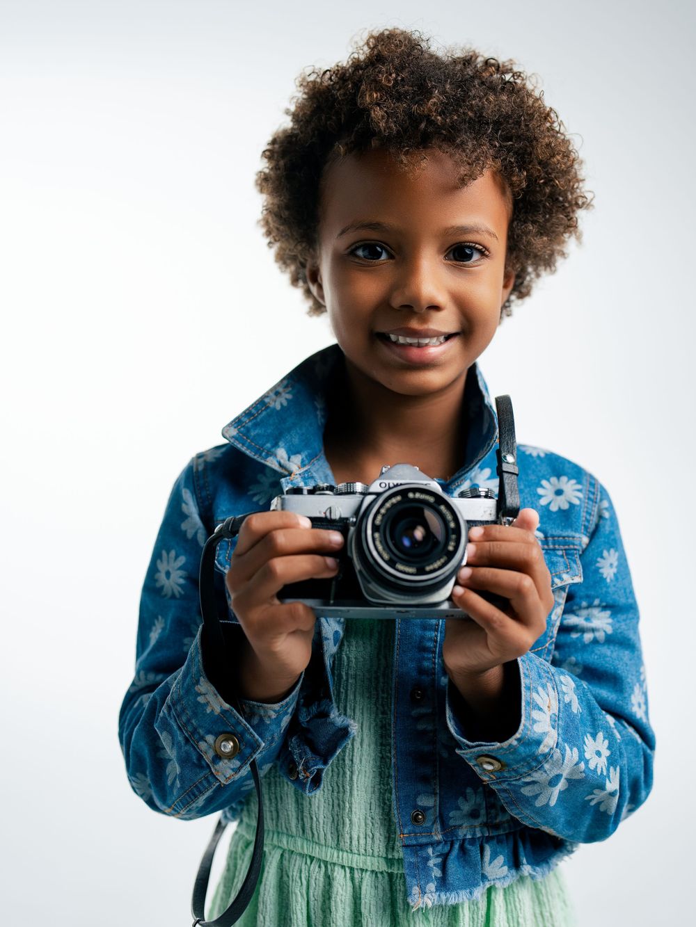 A smiling child with curly hair holds a vintage film camera, wearing a floral denim jacket over a green dress.