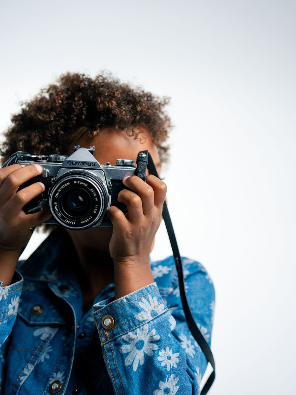 Young person with curly hair holding an Olympus film camera up to their face, wearing a floral denim jacket.