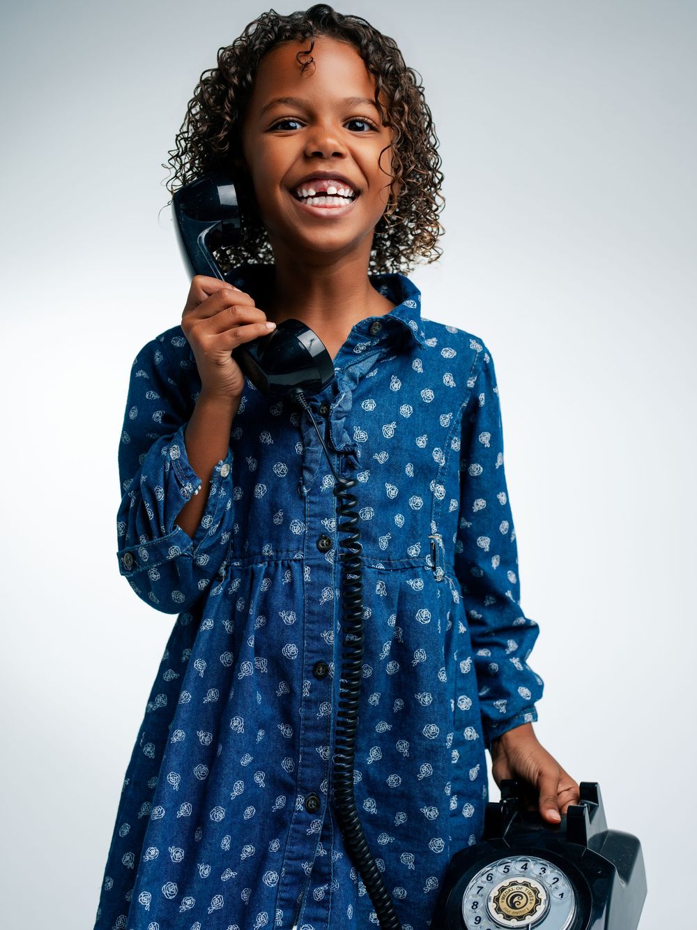 Smiling girl in blue floral dress holding a vintage rotary phone handset, posing against a light gray background.