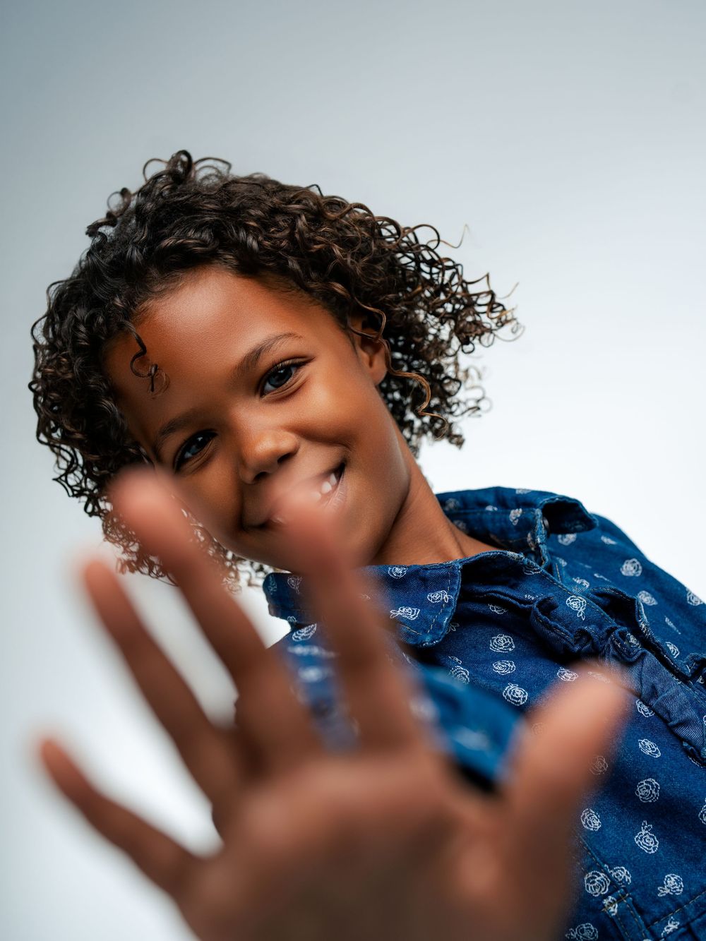 A smiling child with curly hair in a blue shirt reaches toward the camera with an outstretched hand.