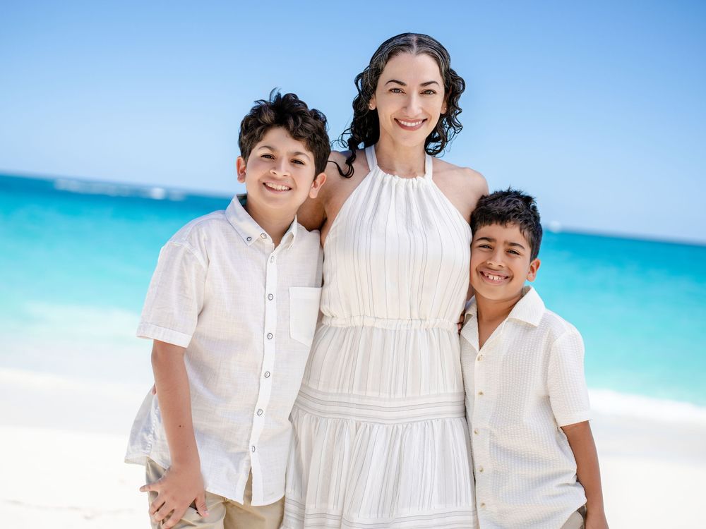 Three people in white outfits pose together on a tropical beach with bright turquoise ocean water in the background.