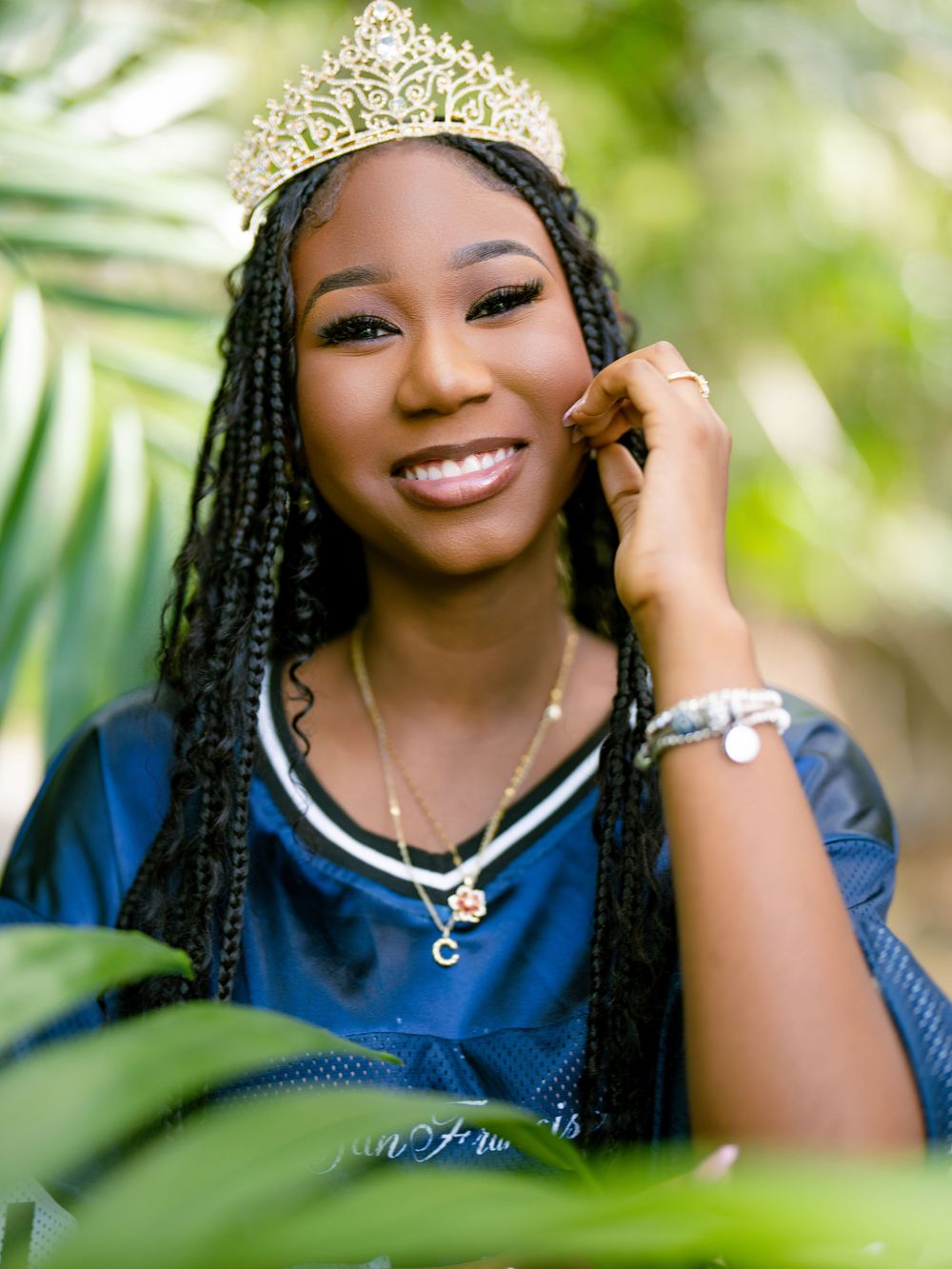 A beaming young person wearing a crown and navy blue top poses elegantly among green tropical foliage outdoors.
