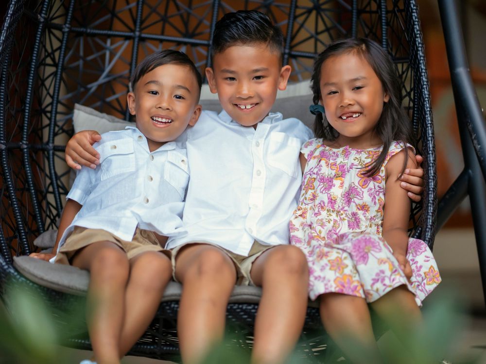 Three children wearing white and floral outfits sit together and smile happily while seated on a bench.