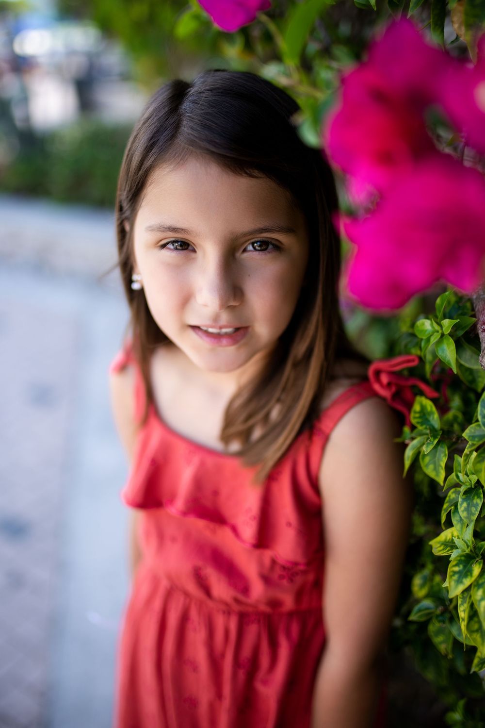 Young girl in red dress smiling beside vibrant pink petunia flowers in an outdoor garden setting.