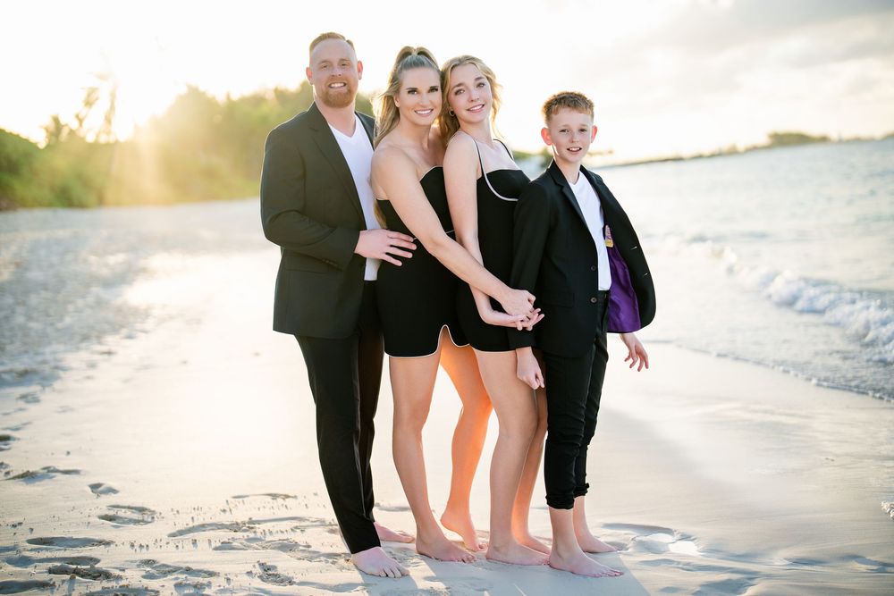 Family of three posing together on a sandy beach at golden hour, wearing elegant black outfits with waves in the background.
