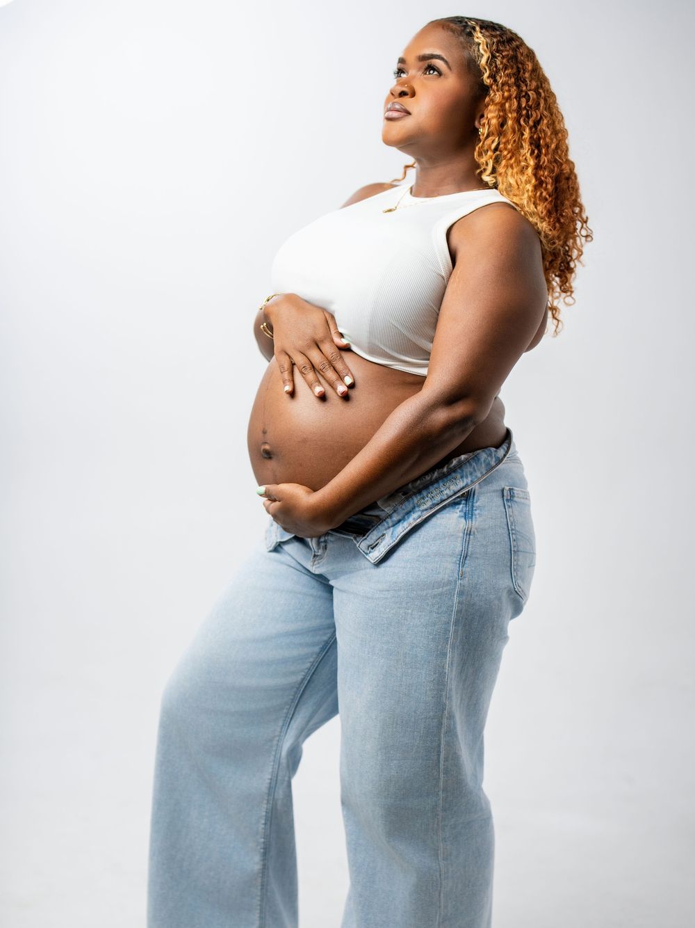 Expecting person in light crop top and wide leg jeans poses against white background while cradling pregnant belly.
