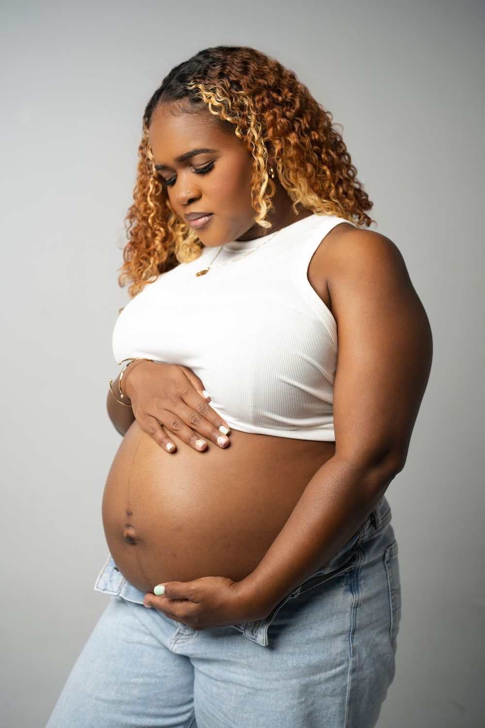 Expectant woman in white crop top and jeans cradles her pregnant belly against a gray background.