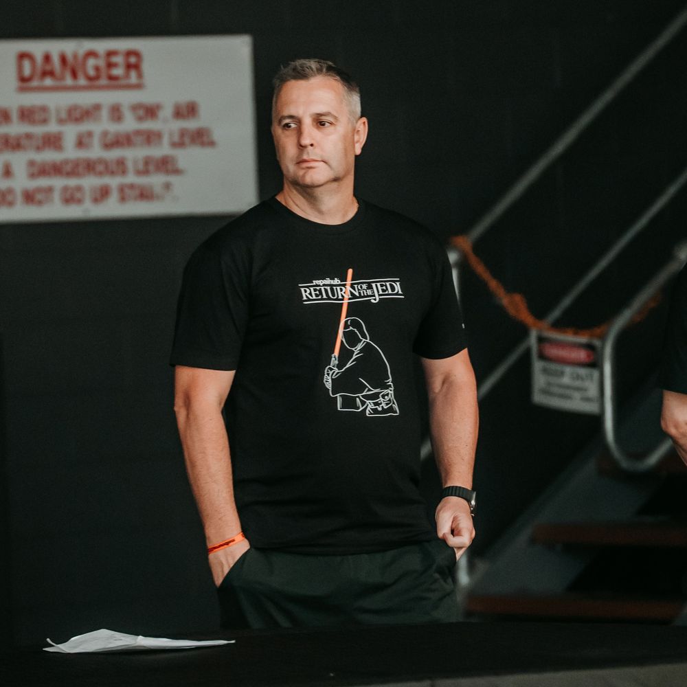 Someone in a black t-shirt sits in a dark room with danger warning signs visible on the wall behind them.
