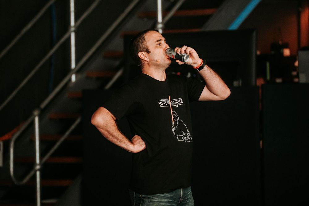 A performer in a black t-shirt energetically sings into a microphone while standing on a dark stage.