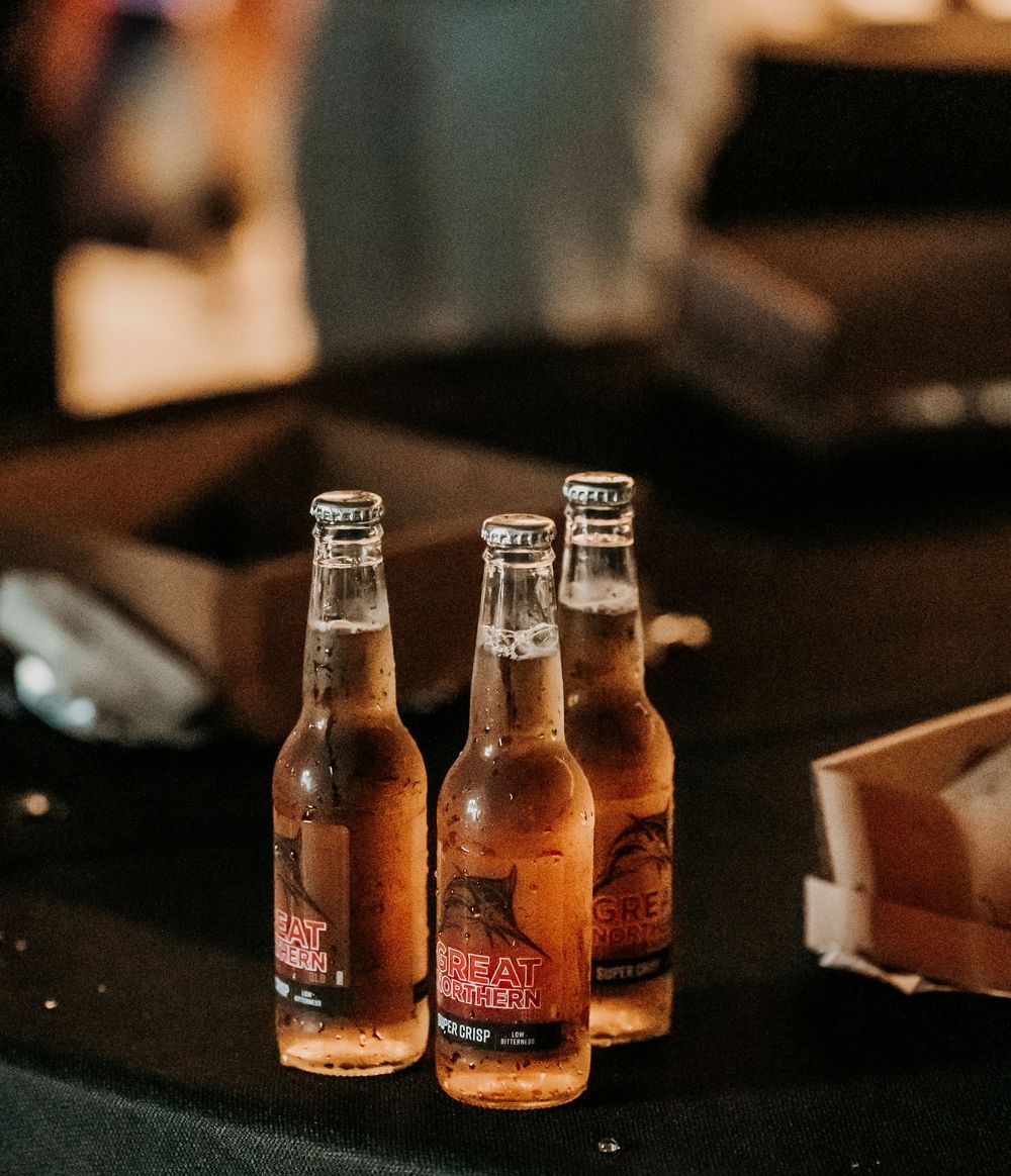 Two glass bottles of beer sit on a dark wooden table with warm moody lighting.