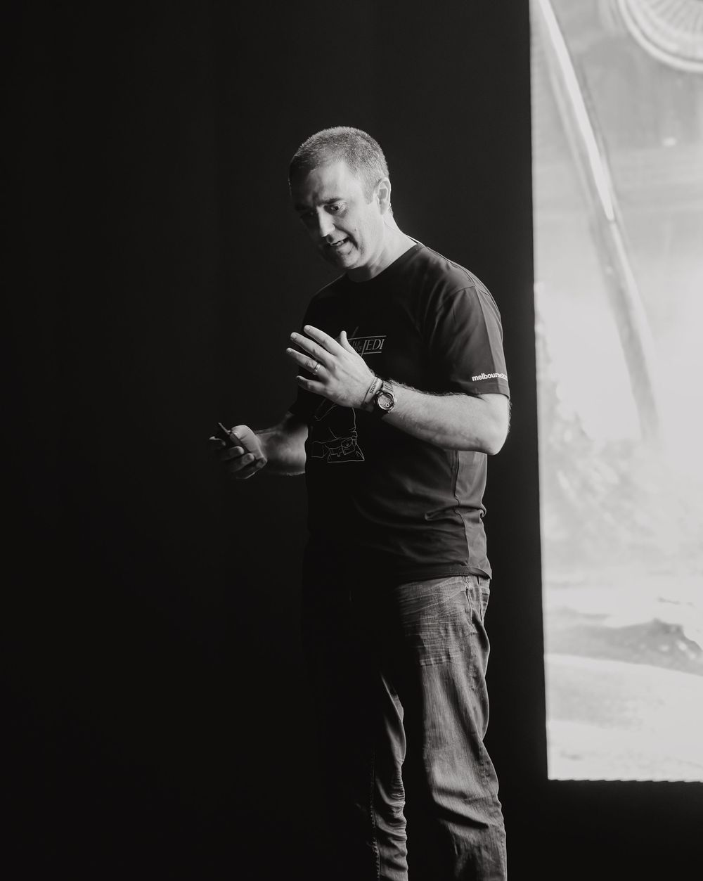 A person in a black t-shirt and jeans stands on stage gesturing with their hands during a presentation.