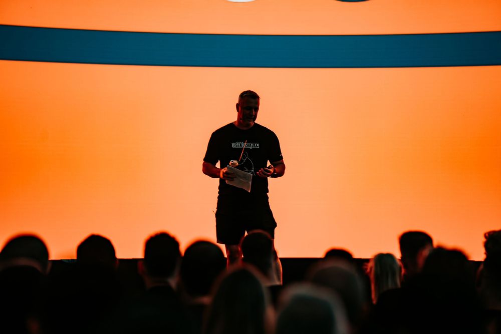 A silhouetted speaker presents on stage against an orange backdrop as an audience watches from darkened seats below.