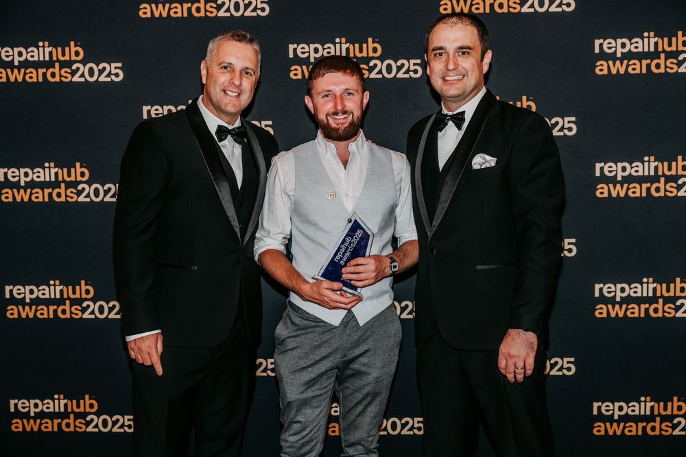 Three people in formal attire posing at an awards ceremony with RepairHub branding visible in the background.