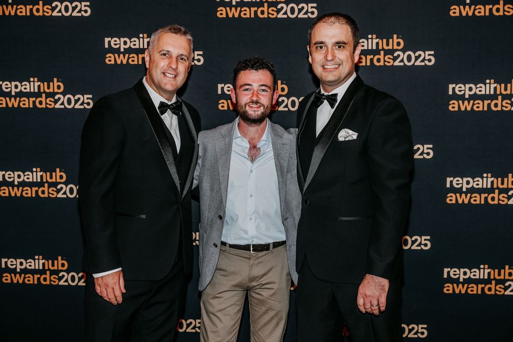 Three people in formal attire pose together at an awards event with a repeating logo backdrop.
