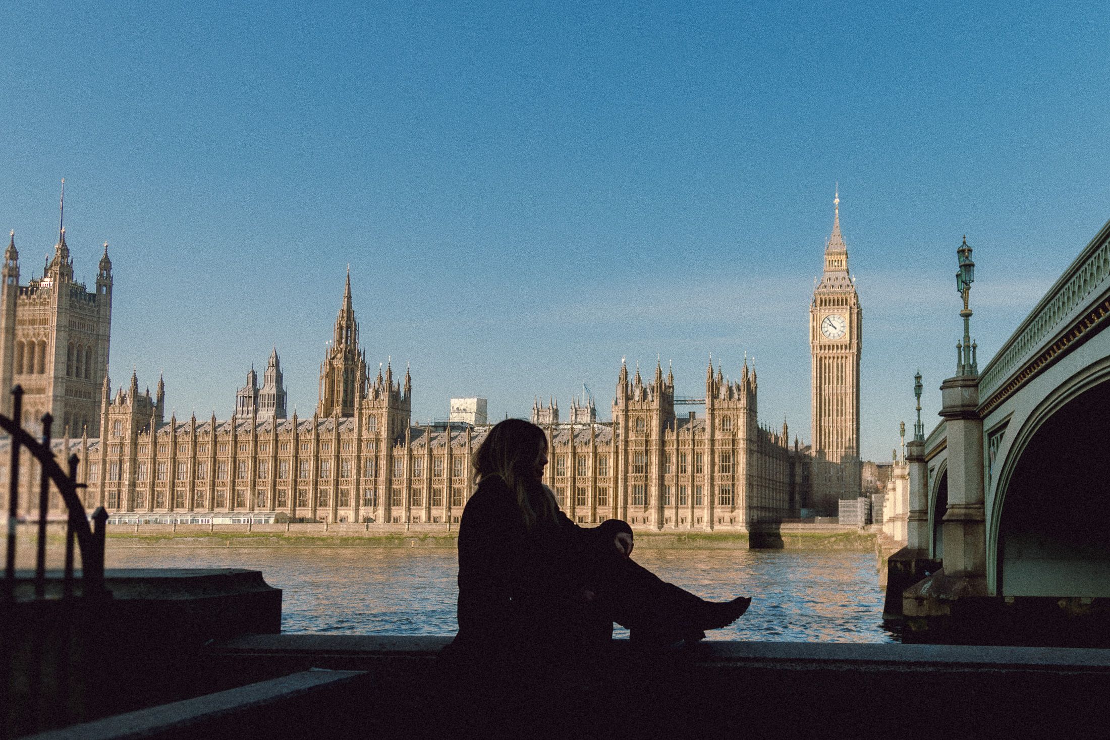 Dark silhouette sits by Thames viewing Houses of Parliament and Big Ben under bright blue sky.