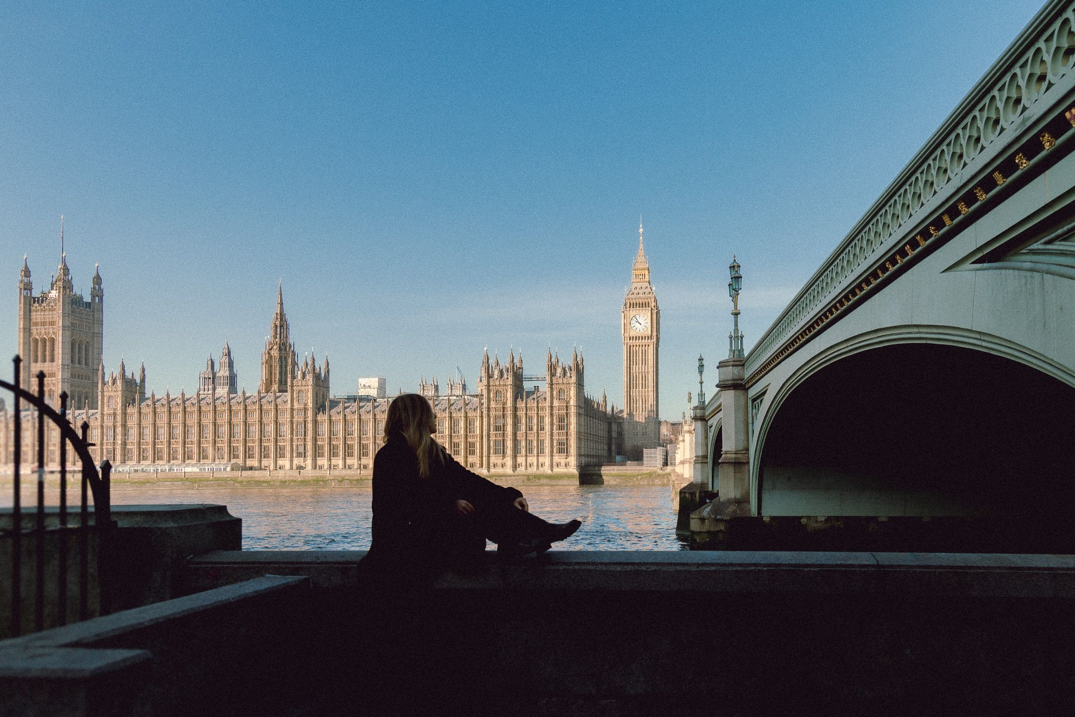 Dark silhouette rests by Thames River with Westminster Palace and Big Ben in sunlight.