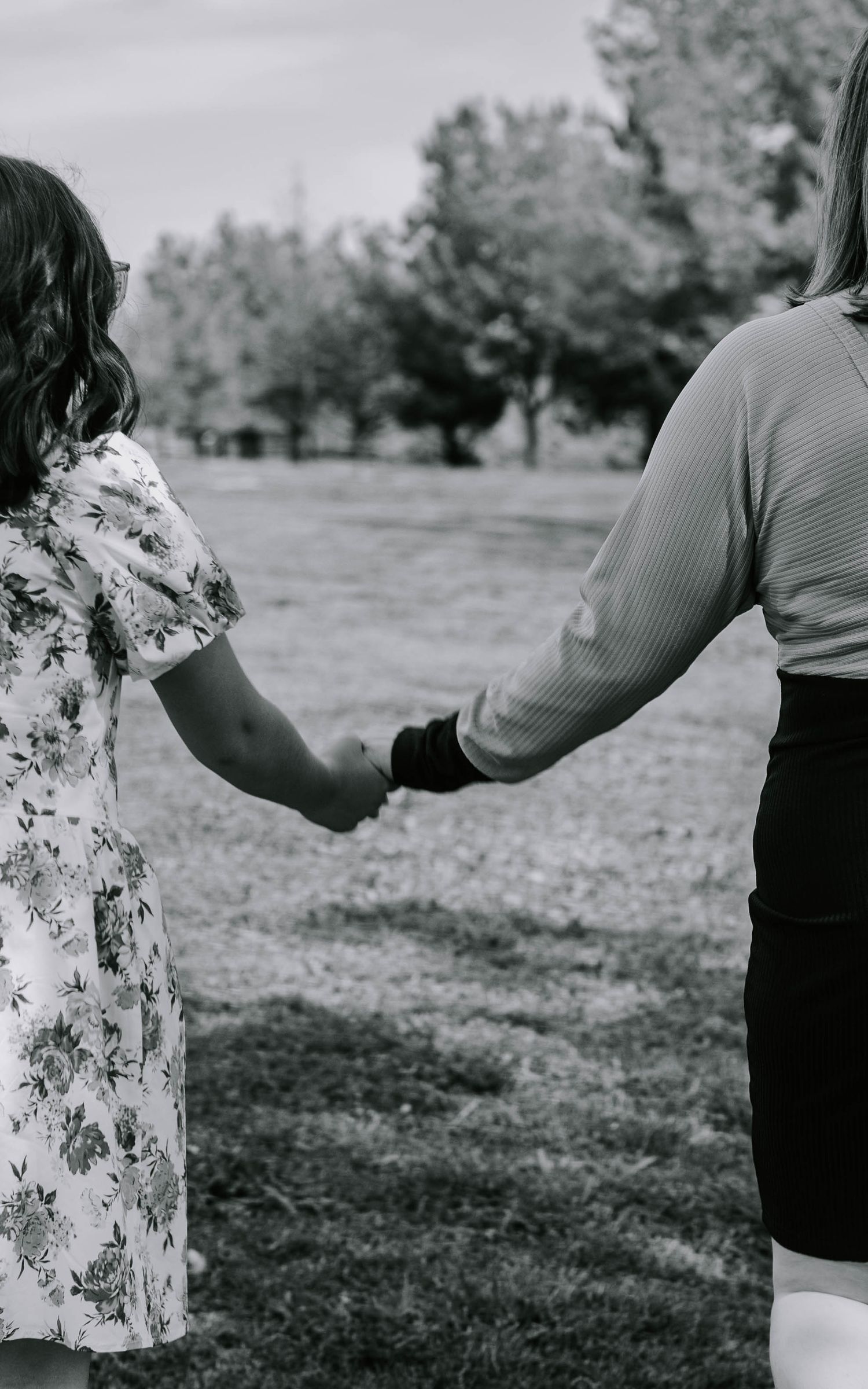 Black and white photo showing two people holding hands while standing in a park with trees in the background.