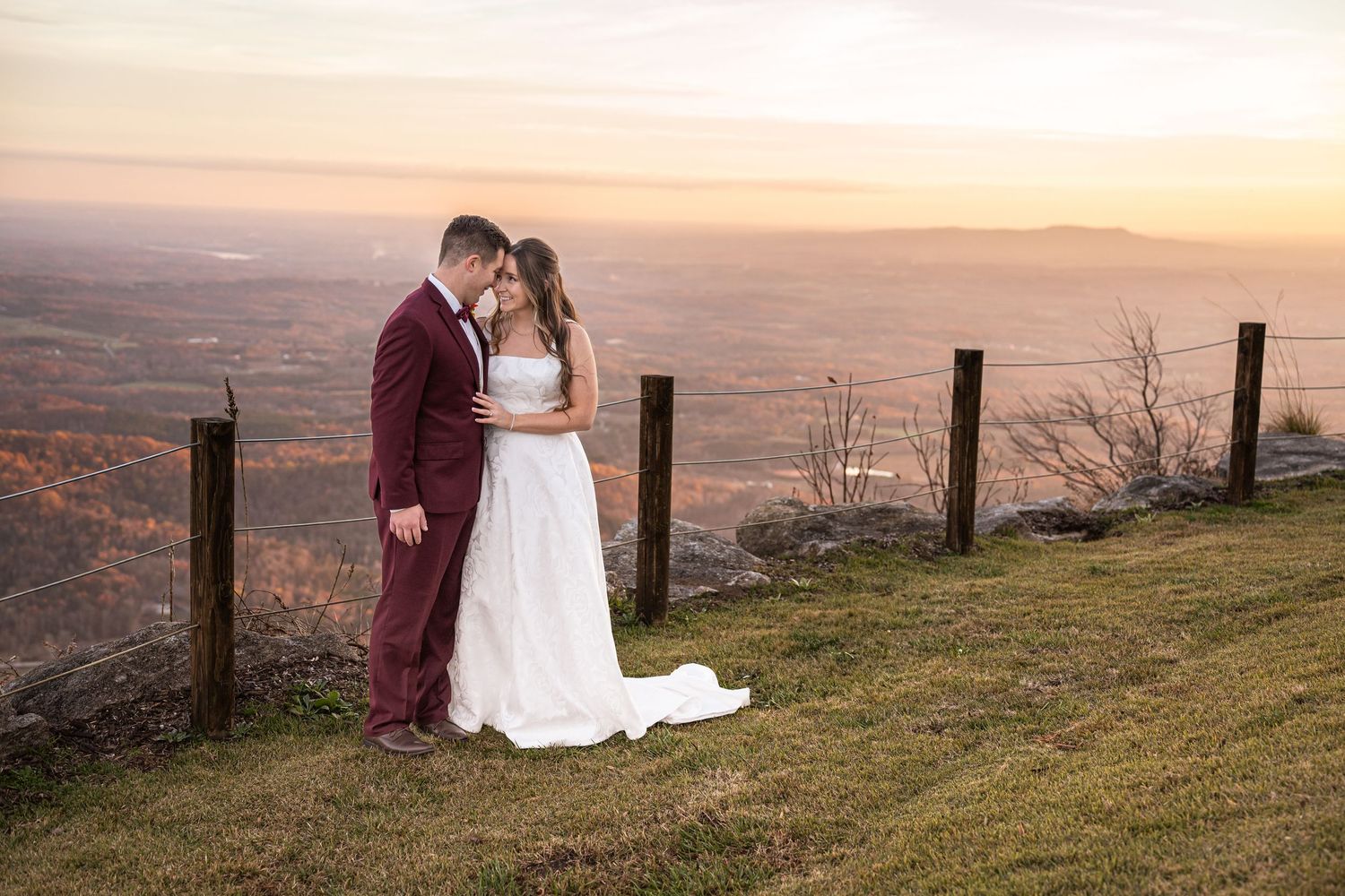 A couple in wedding attire embrace at sunset on a hilltop overlooking a vast autumn valley with wooden fence posts.