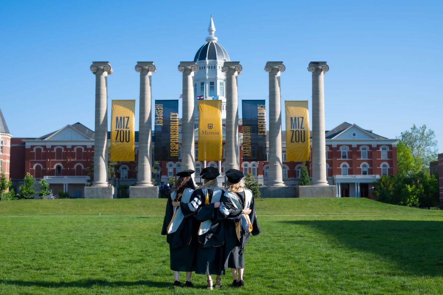 Graduates in black robes celebrate in front of iconic campus columns and architecture.
