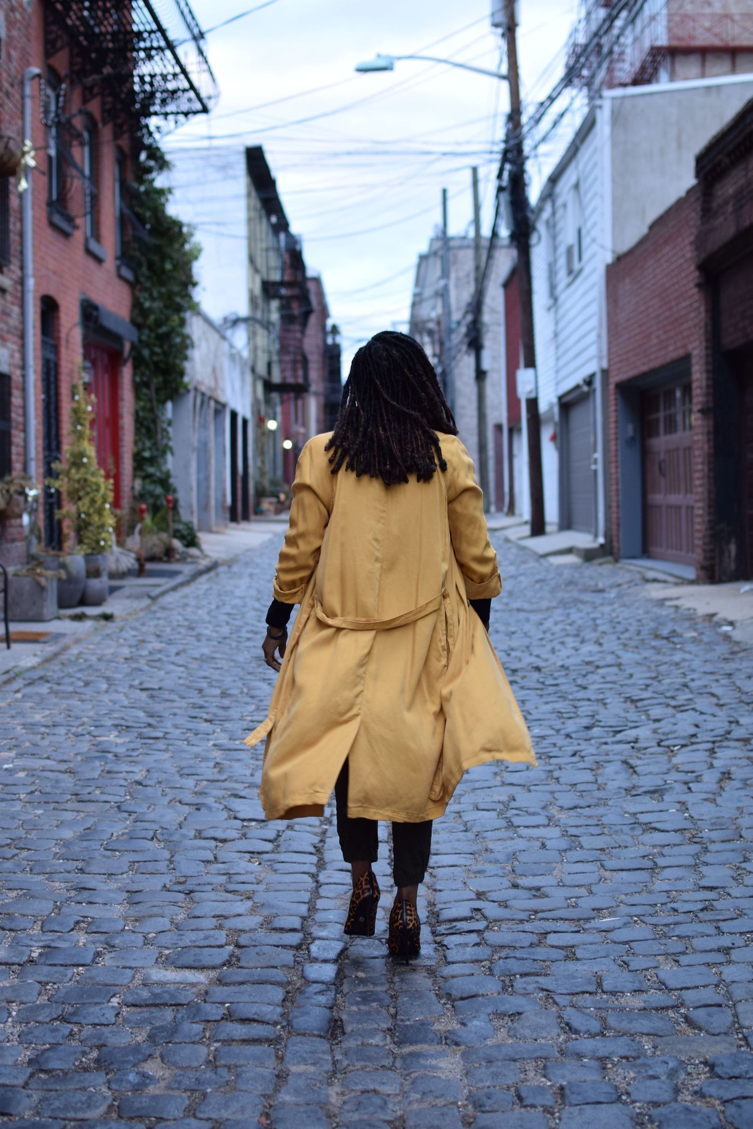 Person wearing a yellow trench coat walks down a cobblestone alley in an urban area on a cloudy day.