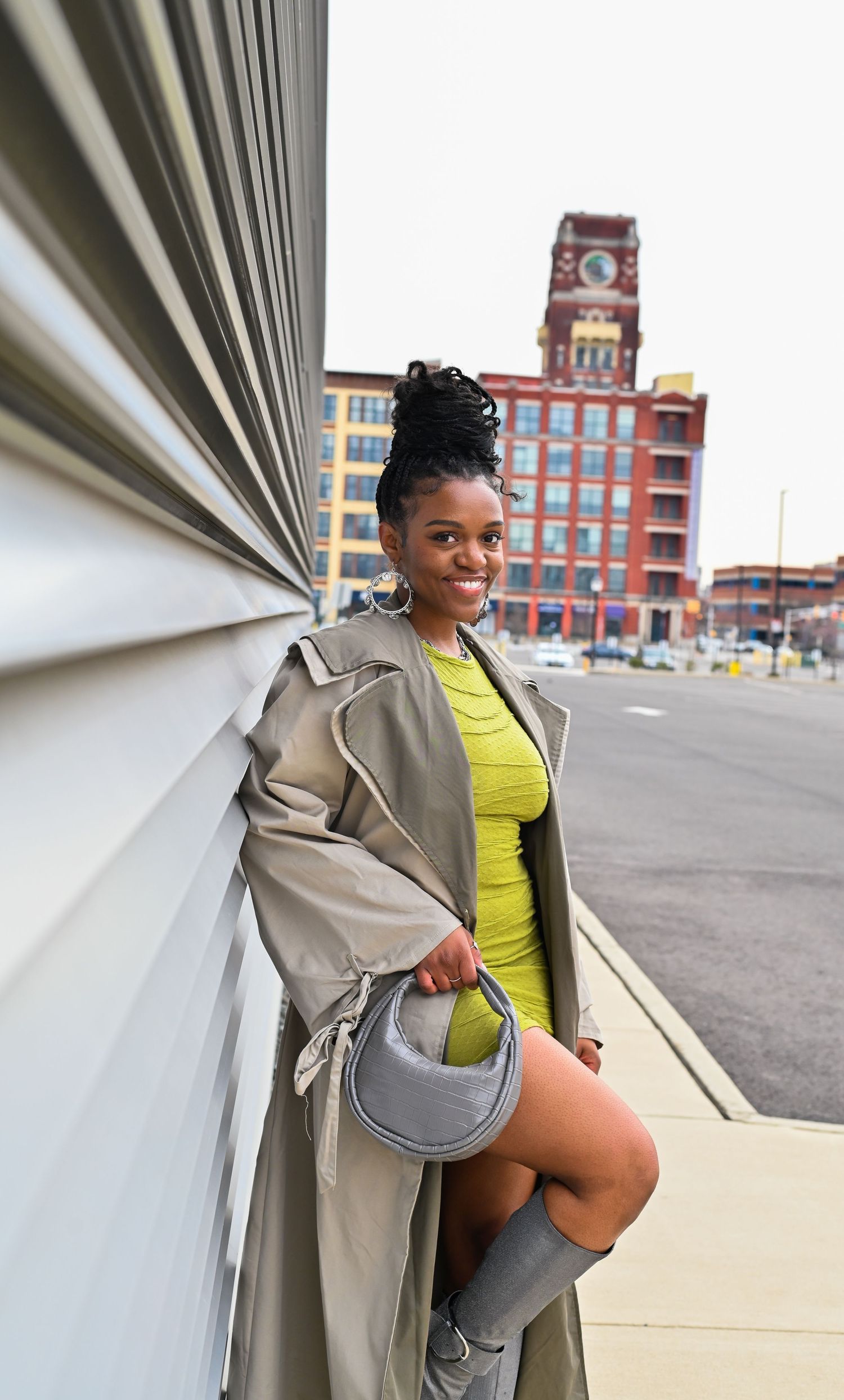 Person in yellow dress and beige trench coat poses confidently against geometric wall with historic brick building in background.