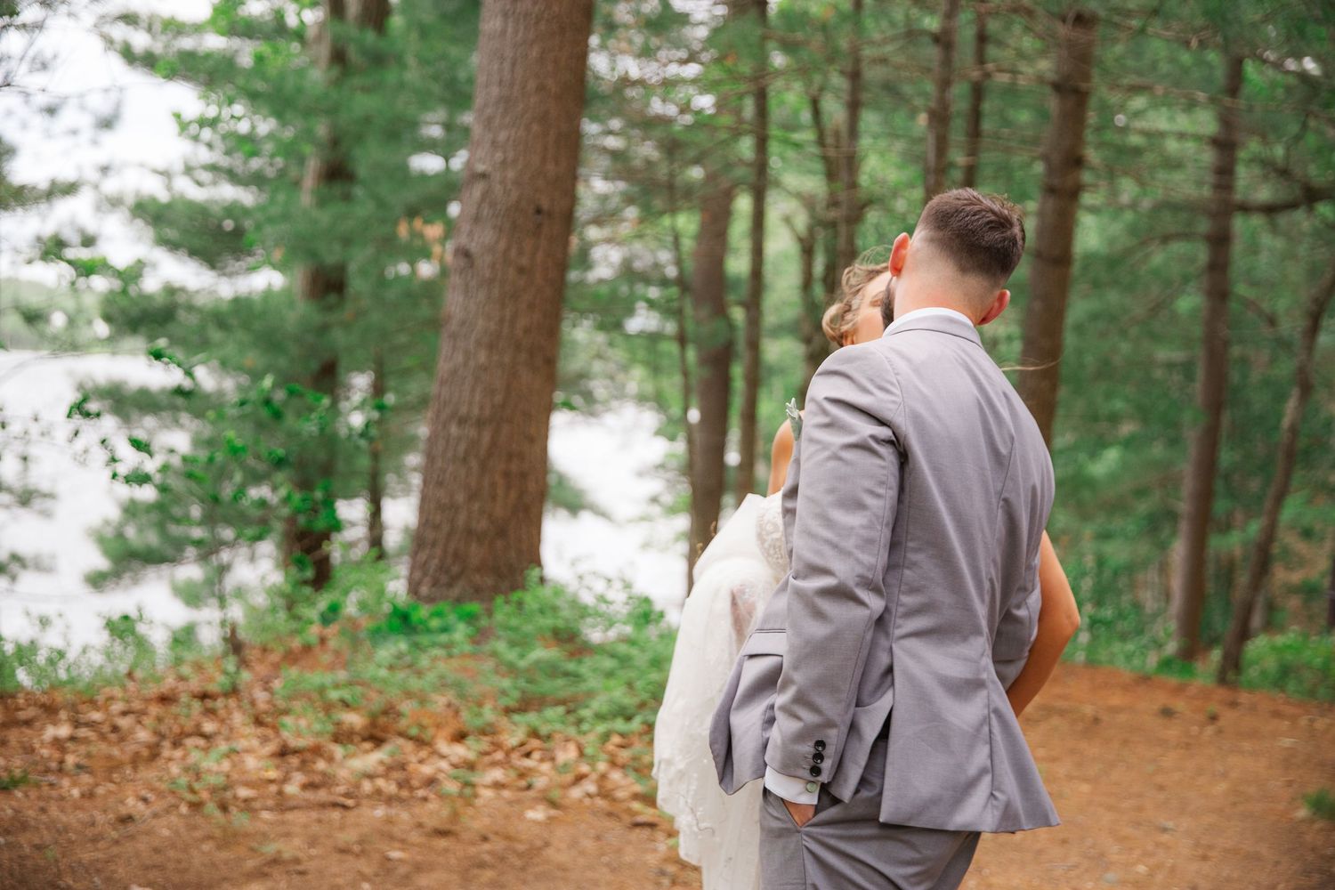 A couple in gray suits walks along a forest path surrounded by tall pine trees and fallen leaves.