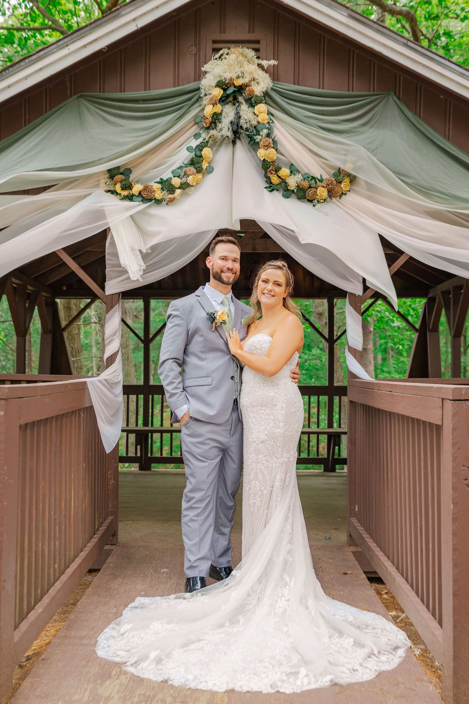A wedding ceremony setup at a wooden gazebo decorated with white fabric draping and floral garlands in a natural setting.