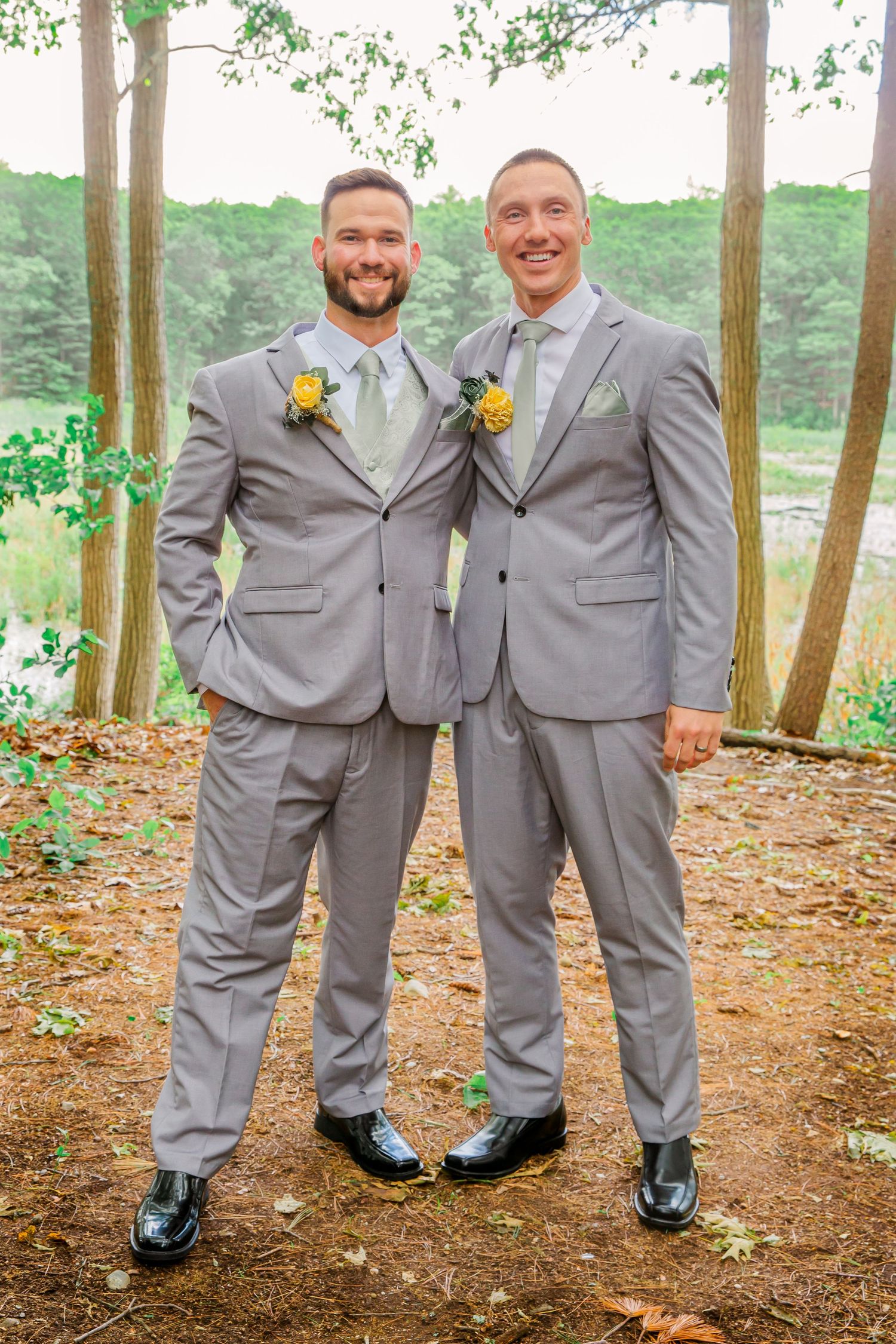 Two people in matching gray suits with yellow boutonnieres stand together in an outdoor wooded setting.