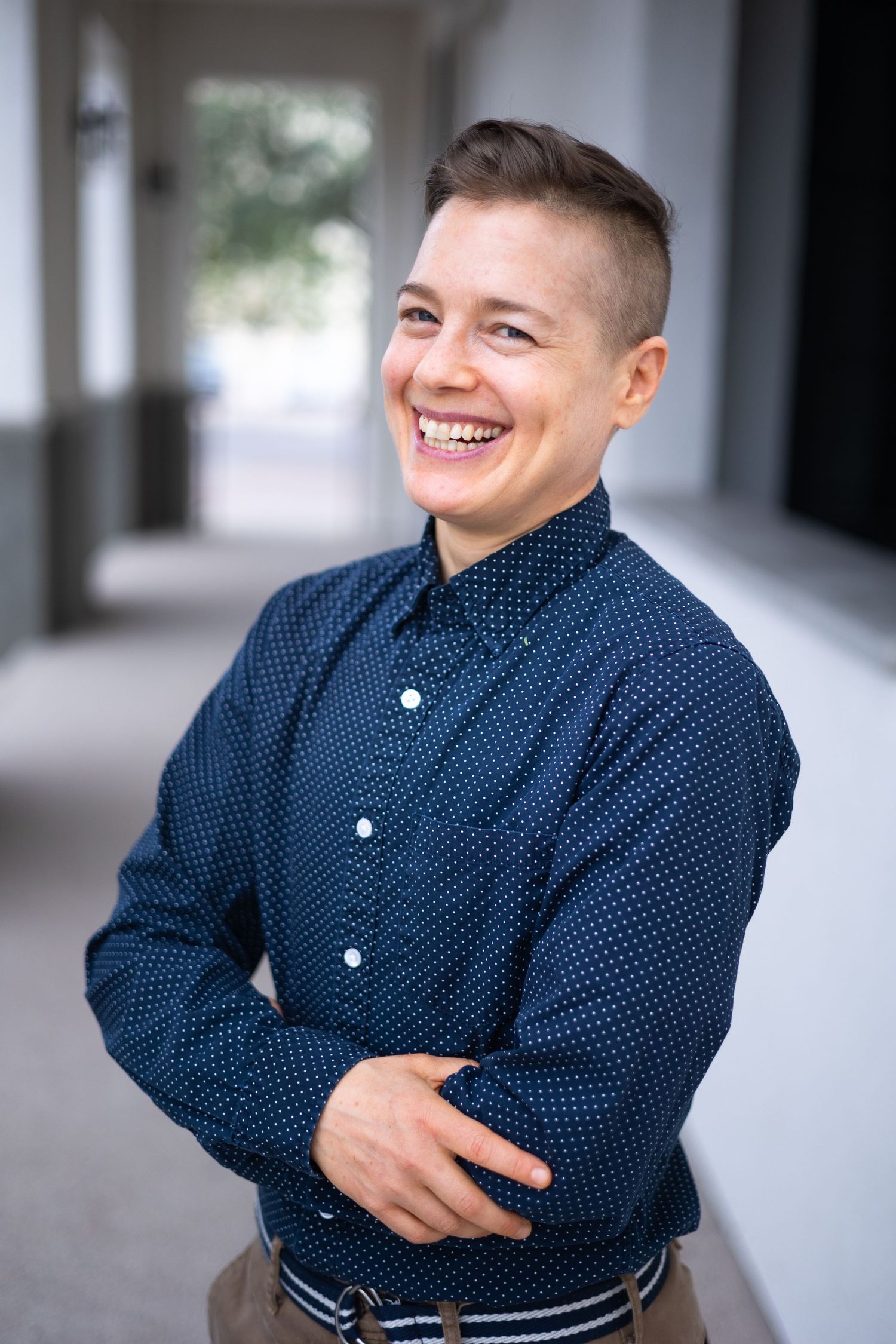 A person wearing a navy polka dot button-up shirt stands in a hallway, smiling warmly for a branding photo in Dallas