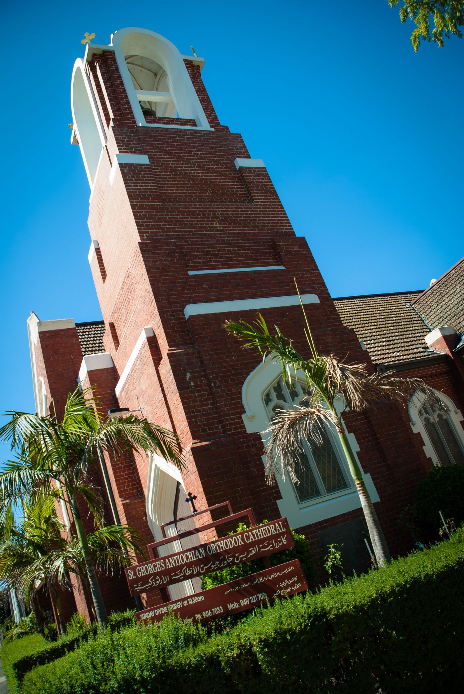 Tall brick church steeple rises against bright blue sky with palm trees and white architectural accents in Southern California.