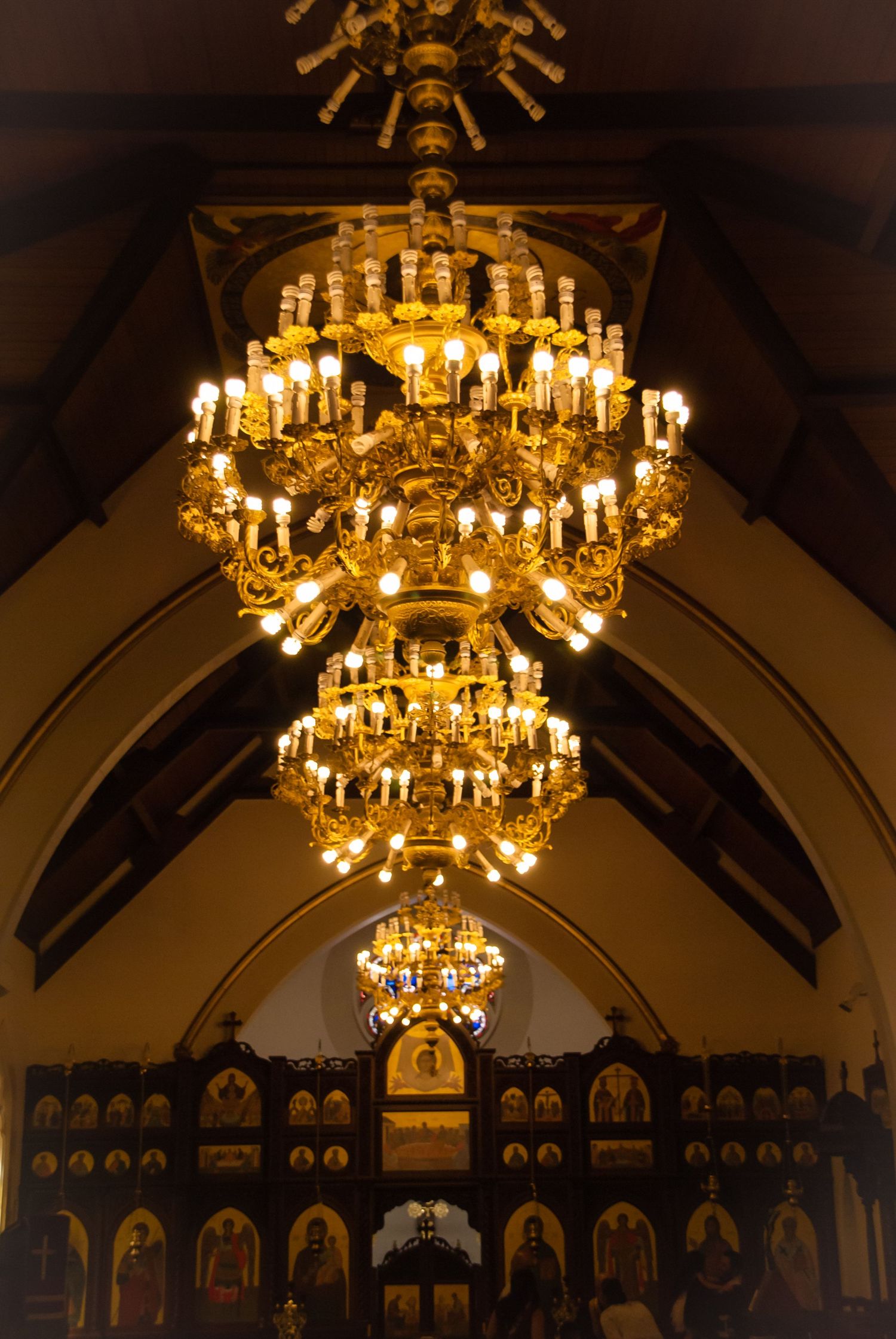 Ornate golden chandeliers illuminate a church interior with arched ceilings and traditional Orthodox iconography.