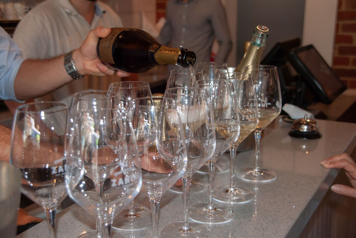 Multiple empty wine glasses lined up on a gray counter with wine bottle being poured in background at tasting event.