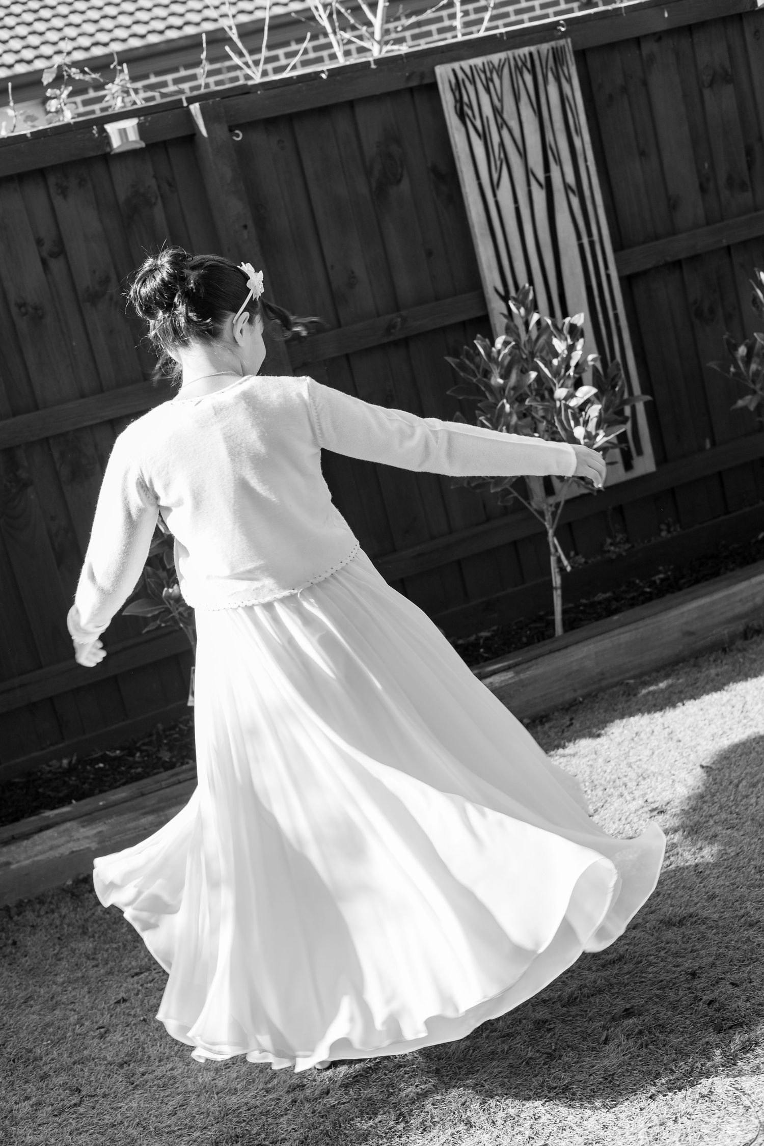 A graceful figure in a flowing white dress twirls in a backyard garden, creating an elegant spinning motion in black and white.
