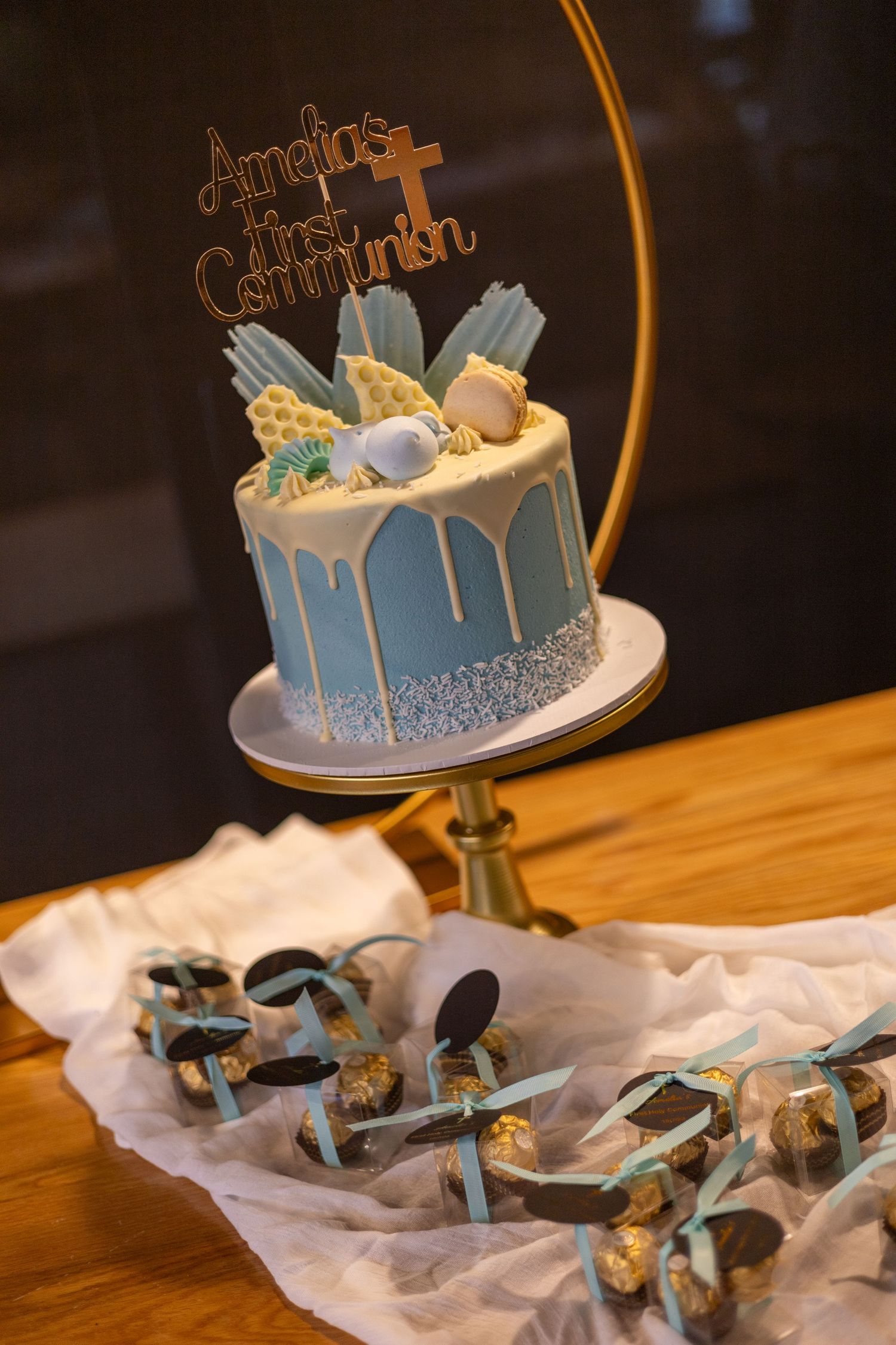 A blue and white communion cake with cross topper and metallic drip decoration sits on a gold stand with party favors below.