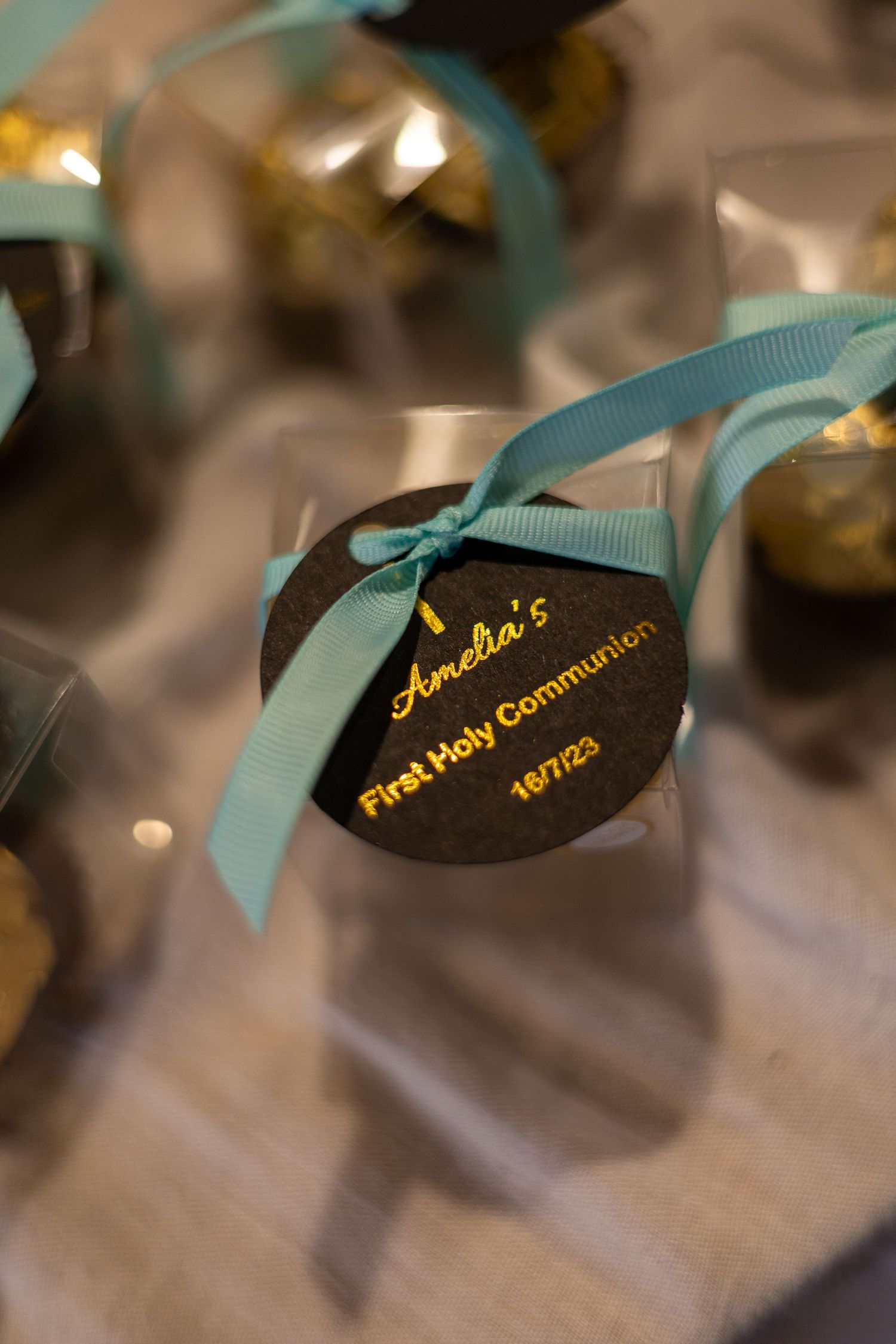 A First Holy Communion favor with a black tag and light blue ribbon features personalized text in gold lettering.